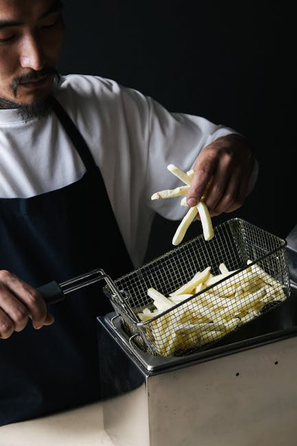 Asian chef placing raw potatoes in deep fryer to make French fries.